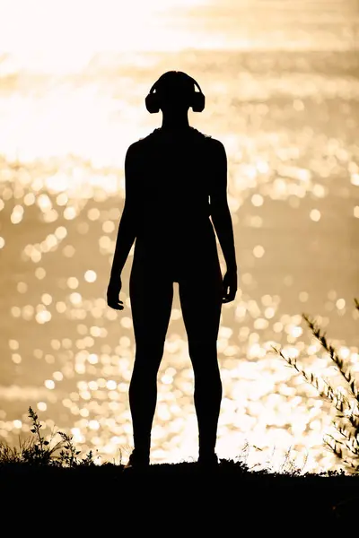 Sporty young woman stands on the shore, lost in music, as sun sets over shimmering water. — Stock Photo