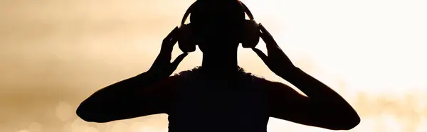 Una mujer atlética hermosa se para en la playa, escuchando música mientras el sol se pone detrás de ella. - foto de stock