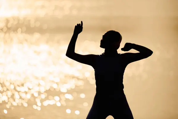 Sporty young woman joyfully dances with confidence by the serene water during sunset. — Stock Photo