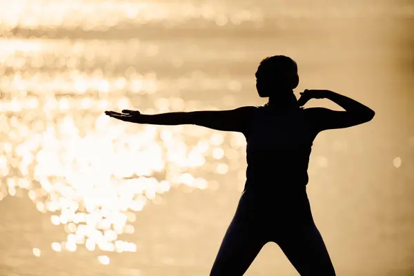 Une jeune femme sportive effectue une pose de yoga alors que le soleil se couche sur l'eau pétillante. — Photo de stock