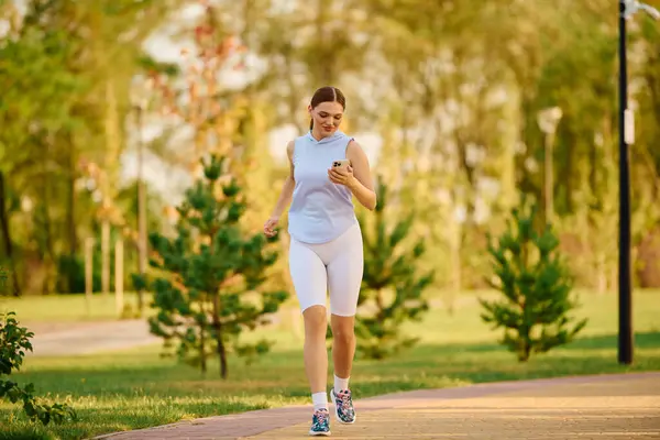 A young beautiful woman jogs through a lush green park, checking her phone while running. — Stock Photo