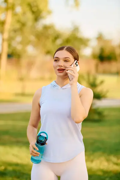 A young woman in athletic wear drinks water while chatting on her phone in a sunny park. — Stock Photo