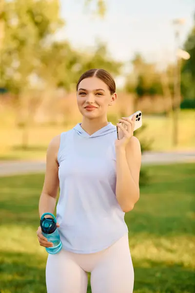 Mujer con una construcción atlética sostiene una botella de agua y un teléfono inteligente en un parque verde soleado. - foto de stock