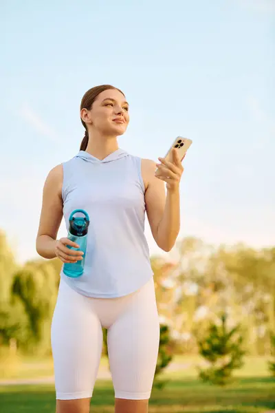 Una mujer atlética sonríe en un parque, revisando su teléfono con una botella de agua en la mano. - foto de stock