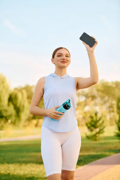 A young woman jogs in a sunny park with a water bottle and smartphone, exuding confidence. — Stock Photo
