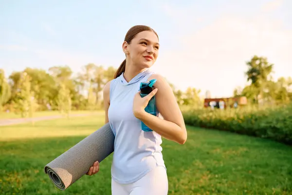 A young, athletic woman walks confidently through a green park, carrying workout gear. — Stock Photo