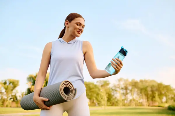 Young beautiful woman enjoys a refreshing moment with a water bottle and yoga mat outdoors. — Stock Photo