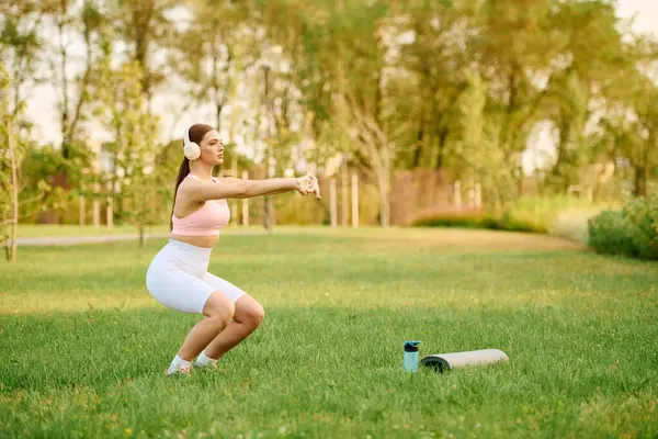 Sporty woman exercises with determination, performing squats on a grassy field surrounded by trees. — Stock Photo