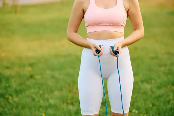 Athletic young woman holds a jump rope, ready to start her workout in the sunlit park. — Stock Photo