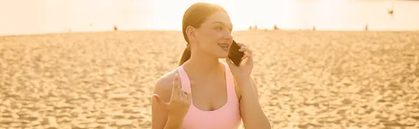 Una joven deportista sonríe por teléfono, disfrutando de la puesta de sol en la playa. - foto de stock