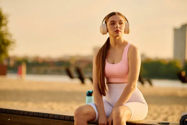 As the sun sets, a beautiful young woman relaxes on a bench, wearing headphones and athletic gear. — Stock Photo