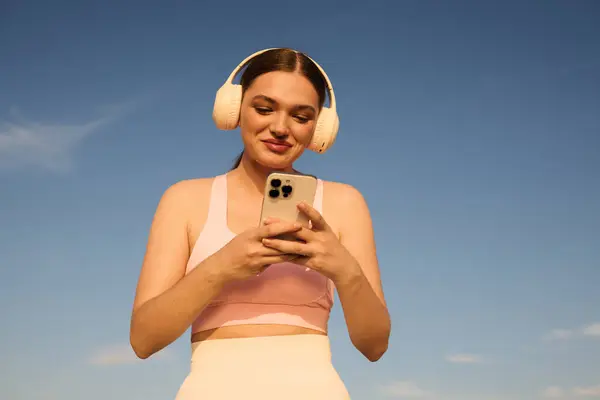 A smiling athletic woman in headphones checks her phone under a blue sky. — Stock Photo