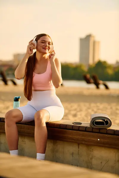 Спортивная женщина улыбается в наушниках, сидя на берегу с бутылкой воды поблизости. — стоковое фото