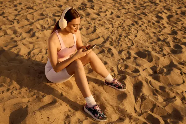 Young athletic woman sits on warm sand, listening to music through headphones while texting. — Stock Photo