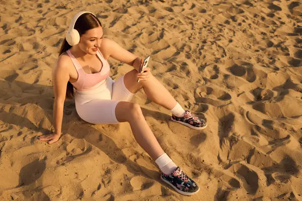 Sporty woman sits on warm sand, smiling at her phone while listening to music. — Stock Photo