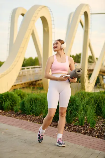 Brightly lit park setting showcases a sporty young woman jogging, headphones on, ready for yoga. — Stock Photo