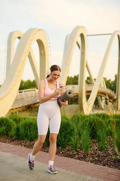 Sporty woman walks with yoga mat, checking her phone near an artistic bridge in the park. — Stock Photo