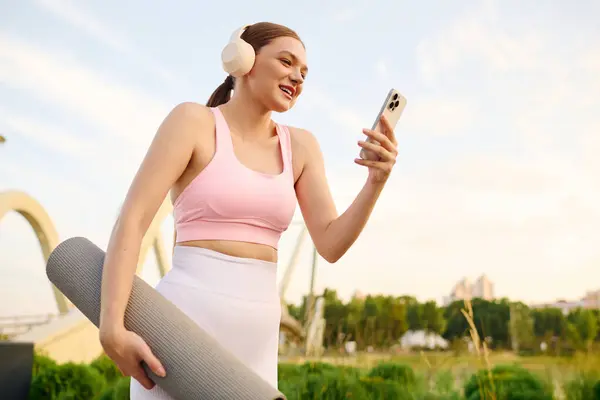 Sporty woman wearing headphones smiles while checking her phone in a vibrant outdoor setting. — Stock Photo
