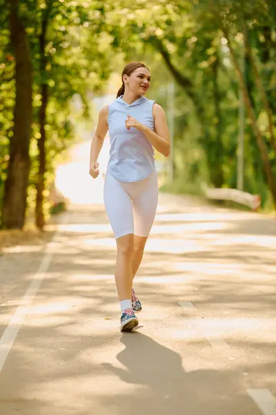 Giovane donna sportiva corre con un sorriso su un bellissimo sentiero alberato alla luce del sole. — Foto stock