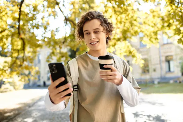 Un jeune homme aux cheveux bouclés se tient debout dans un cadre lumineux, tenant un café et regardant son téléphone. — Photo de stock