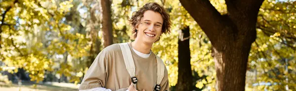 Young man with curly hair smiles brightly while standing under a tree on a sunny day. — Stock Photo