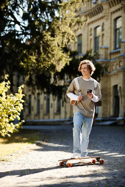 A young man skates down a sunny path, smiling and checking his phone. — Stock Photo
