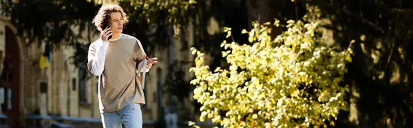 A stylish young man walks through an urban area, casually using his smartphone among greenery. — Stock Photo