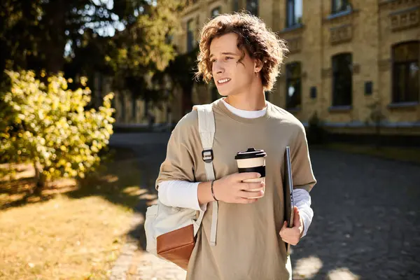 A stylish young man walks on campus, holding a warm drink and laptop, smiling in autumn. — Stock Photo