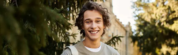 Handsome young man smiles brightly while standing amid trees near an educational institution. — Stock Photo