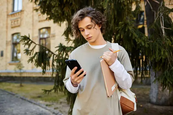 Un joven con estilo en ropa casual comprueba su teléfono, sosteniendo un cuaderno y un lápiz en la vegetación. - foto de stock