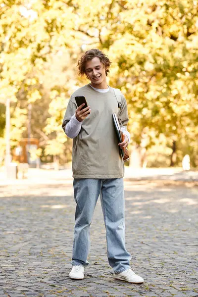 A stylish young man smiles at his phone in a colorful park as leaves fall around him. — Stock Photo
