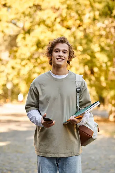Giovane studente cammina fiducioso lungo un sentiero alberato, tenendo libri e un telefono, sorridendo. — Foto stock