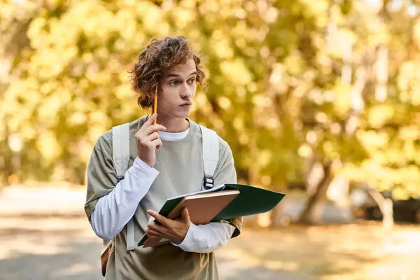 A young man with stylish hair holds notebooks and a pencil, deep in thought among golden trees. — Stock Photo