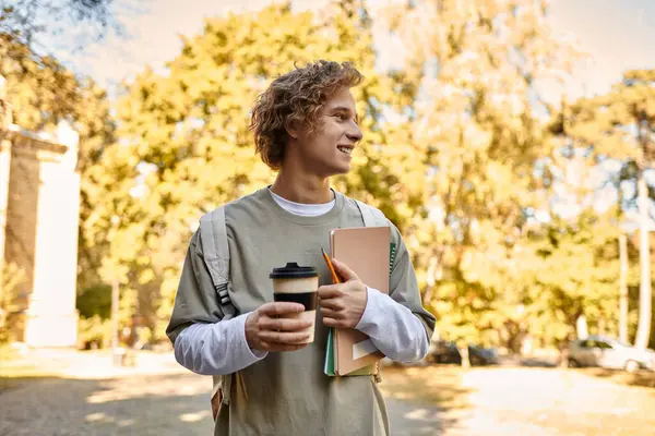 Joven sonríe brillantemente mientras sostiene una taza de café y libros, rodeado de follaje otoñal. - foto de stock