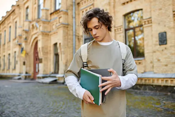 Handsome man with curly hair holds notebooks while standing outside a historic campus building. — Stock Photo