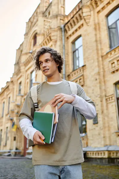 Jovem encantador caminha por uma rua histórica, carregando cadernos e sorrindo para os transeuntes. — Fotografia de Stock