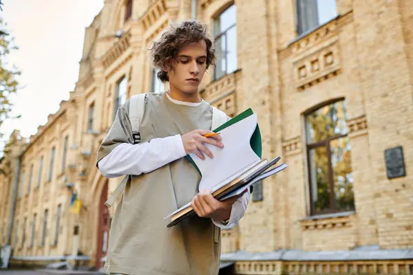 A young man in stylish casual wear reviews teaching materials by an old brick university. — Stock Photo