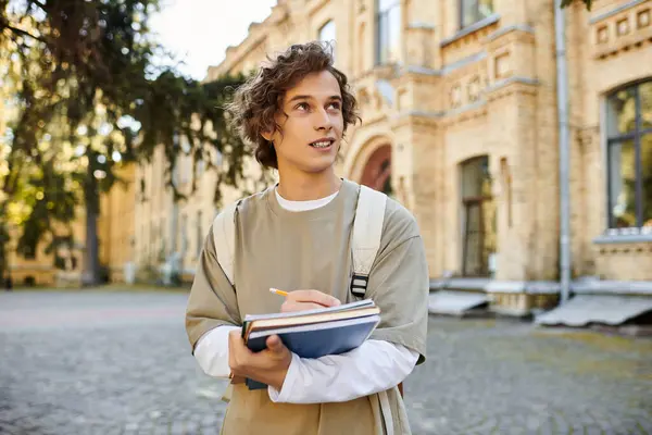 Un giovane uomo elegante con i capelli ricci sta fuori, tenendo quaderni e una matita, pensieroso. — Foto stock