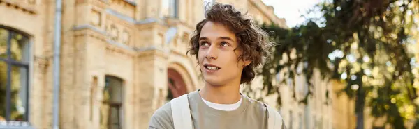 A vibrant young man with curly hair smiles while exploring his university campus on a bright day. — Stock Photo