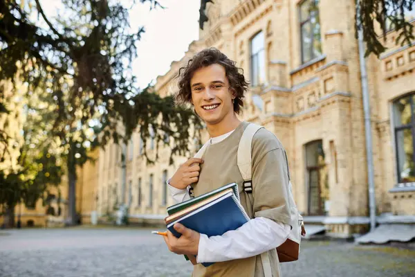 Un joven sonríe, sosteniendo libros mientras pasea por un campus encantador. - foto de stock