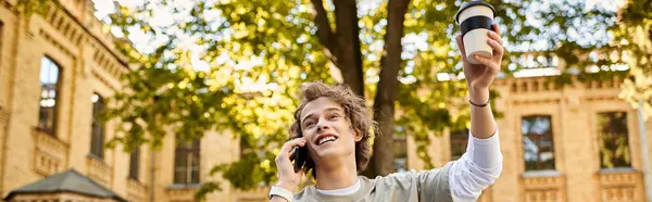 Stylish student joyfully talks on the phone with a drink in hand amidst a vibrant campus setting. — Stock Photo