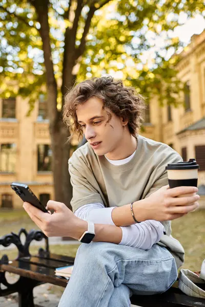 Joven con el pelo rizado se relaja en un banco al aire libre, sosteniendo una taza de café y usando su teléfono. - foto de stock