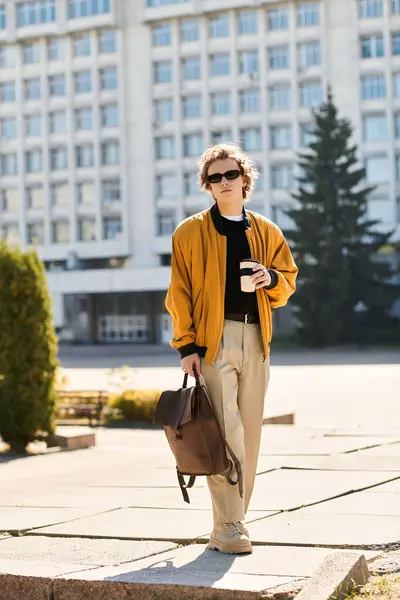 A young stylish man strolls across a university campus carrying coffee and a brown backpack. — Stock Photo