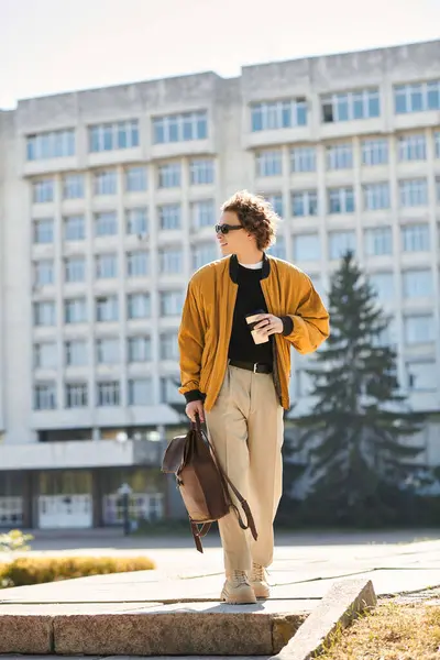 Handsome man dressed in trendy attire walks casually with coffee and backpack on a bright day. — Stock Photo