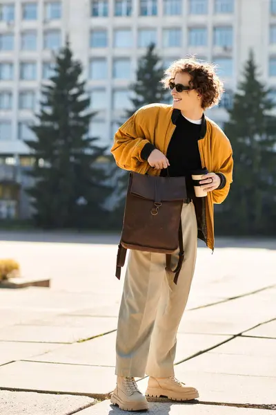 Charming young man dressed in trendy attire holds a coffee cup and a backpack in a sunny plaza. — Stock Photo