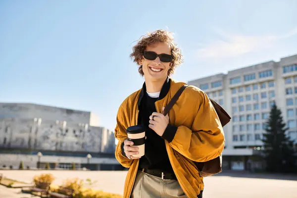 A confident young man with stylish attire holds a cup of coffee and smiles brightly while walking. — Stock Photo