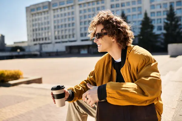 Young man with curly hair and sunglasses sits on steps, savoring his coffee in a sunny city plaza. — Stock Photo