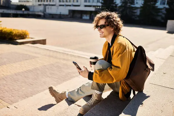 A fashionable young man sits on the steps, sipping coffee and smiling as he uses his smartphone. — Stock Photo
