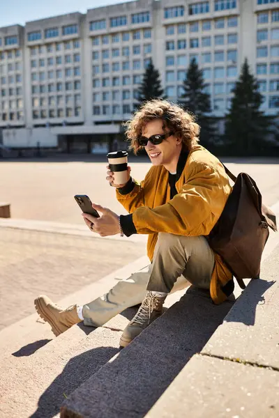 Un giovane uomo elegante siede su un gradino, sorridendo mentre si gode il caffè e usando il suo smartphone. — Stock Photo