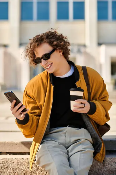 Trendy young man lounges outdoors, sipping coffee and checking his phone in the sun. — Stock Photo
